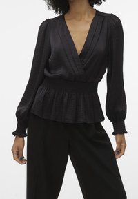 Blouse noire à manches bouffantes longues, en tissu texturé, avec une taille smockée. Dotée d'un décolleté en V profond et d'un ourlet peplum évasé.