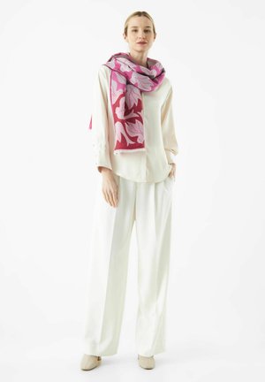 Vrouw in een crèmekleurige blouse en wijde broek, met een roze en rode bloemige sjaal, staand met handen in de zakken op een witte achtergrond.