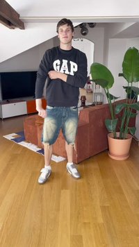 Sweatshirt preto com o logótipo branco "GAP", calções de ganga desgastados e ténis prateados. Ambiente interior com plantas e um sofá castanho.