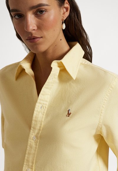 Dzeltena kokvilnas krekls ar klasisku apkakli, ar izšūtu logotipu kreisajā krūtīs un ar pogu aizdari priekšpusē.
