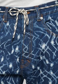 Džinsai su mėlyna ir balta abstrakia raštais, su traukimo uždarymu, oranžiniais siūlų pasiūlymais ir dėvėtais akcentais.