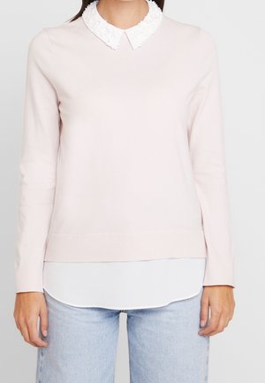 Mujer con suéter rosa claro de manga larga con cuello blanco con volantes y bajo de camisa blanco, combinado con vaqueros azul claro.
