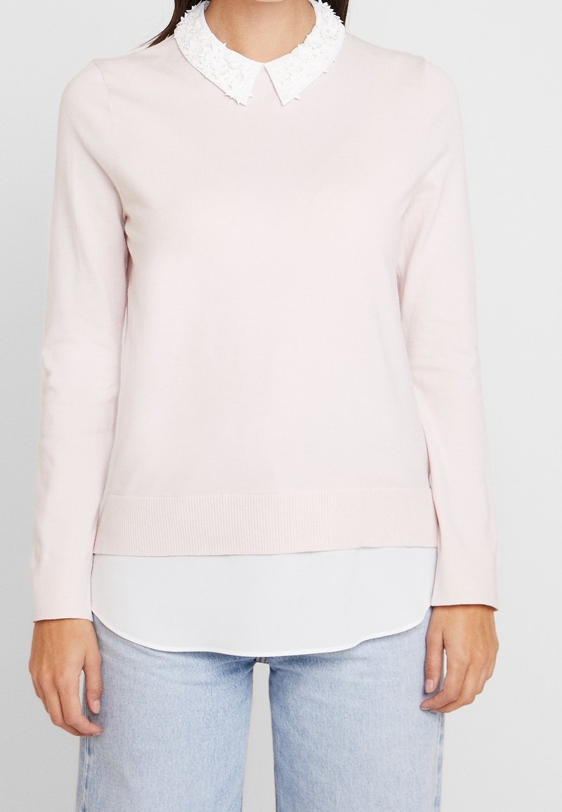 Femme portant un pull rose clair à manches longues avec un col à volants blanc et un bas de chemise blanc, associé à un jean bleu clair.