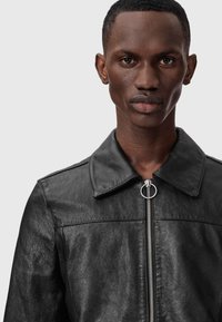 Close-up of a young man with short hair wearing a black leather jacket with a silver zipper and round pull tab, looking straight ahead.