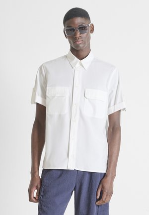 Man draagt een oversized wit kort mouwen overhemd met knopen en voorzakken, en donkere zonnebril, gecombineerd met gestructureerde blauwe broek.