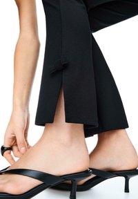 Svarta cropped byxor med sidoflikar och rosett på nederkanten, tillsammans med svarta högklackade sandaler med remmar. Slät textur.