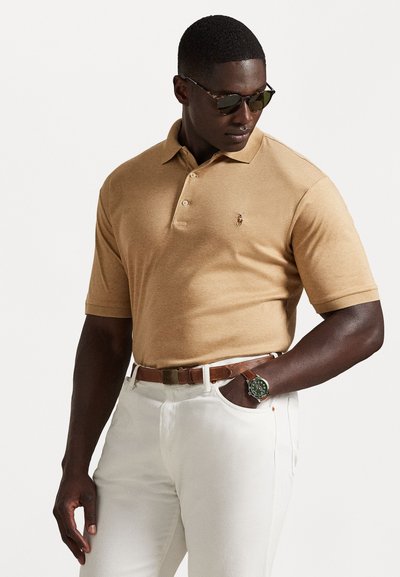 Kortærmet, beige polo-skjorte med tre knapper, broderet logo på brystet, parret med hvide bukser og et brunt læderbælte. Grøn ur på håndleddet.