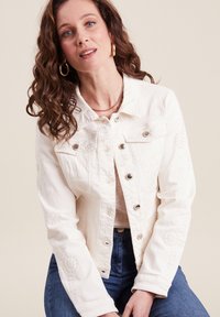 Witte denimjack met bloemenborduursel, klassieke kraag, knoopsluiting en een getailleerd ontwerp. Gecombineerd met blauwe jeans voor contrast.