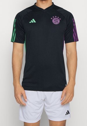 Mann trägt schwarz FC Bayern München Fußballtrikot mit grünen und violetten Streifen und weißen Adidas-Shorts, steht vor weißem Hintergrund.
