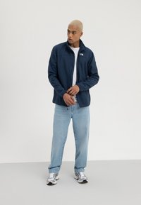 Jachetă zip-up albastră închisă cu guler înalt, confecționată dintr-un material moale. Asortată cu blugi de un albastru deschis și pantofi sport argintii.