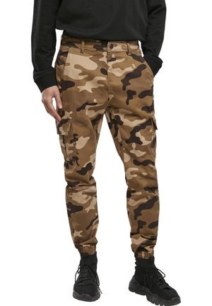Bărbat purtând pantaloni cargo de camuflaj maro și bej cu manșete elastice, pantofi negri și un top negru cu mâneci lungi, cu mâinile în buzunare.