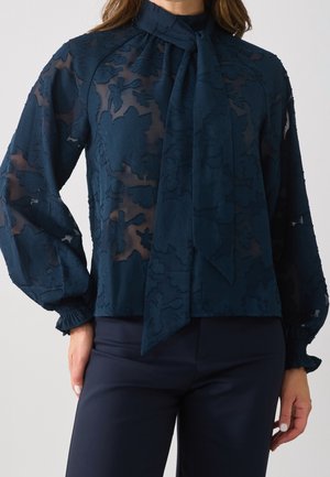 Vrouw draagt een marineblauw doorschijnend bloemenblouse met een grote strik bij de hals en lange opgeblazen mouwen, gecombineerd met marineblauwe broek.