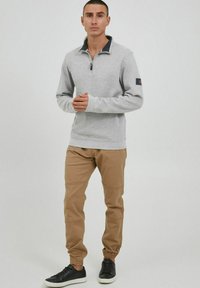 Sivi zip-up pulover s mornarskom ovratnikom, uparen s bež hlačama za trčanje koje imaju panel na koljenima i uske gležnjeve. Crne tenisice upotpunjuju izgled.