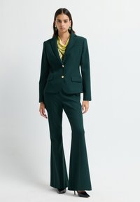 Mujer vestida con un blazer entallado verde oscuro y pantalones acampanados, con una blusa amarilla-verde de estampado y tacones negros, de pie contra un fondo liso.