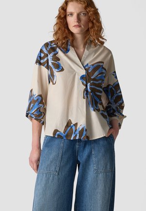 Woman with red curly hair wears beige blouse with large blue and brown floral prints and loose blue denim jeans, standing against plain background.