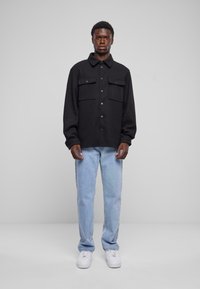 Zwarte overhemd met knoopsluiting, gemaakt van een textuurstof, met twee borstzakken; gedragen met lichtblauwe jeans en witte sneakers.