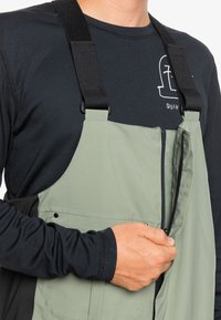 Salopette étanche verte avec des bretelles noires réglables, fermeture éclair et une poche latérale, portée par-dessus une chemise noire à manches longues.