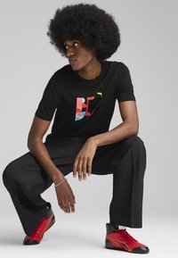 Sort t-shirt med korte ærmer og farverigt grafisk design. Modellen bærer løstsiddende sorte bukser og røde højhælede sko med sorte accenter.