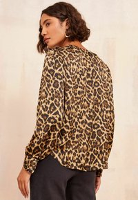 Leopardenmuster-Bluse aus leichtem Stoff; mit langen Ärmeln, abgerundetem Saum und knöpfbaren Manschetten; Farben umfassen Beige, Schwarz und Braun.