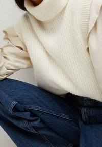 Pull en maille couleur crème avec col roulé et texture côtelée, superposé à une blouse légère et ondulée. Associé à un jean en denim bleu.