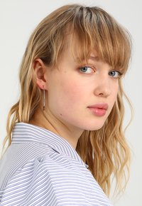 De longues boucles d'oreilles argentées au design élégant et droit. Le modèle a des cheveux ondulés châtain clair et porte une chemise à col rayée.