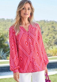 Overhemd met een roze en rode geometrische diamantenprint, lange mouwen met elastische manchetten en een relaxte pasvorm. Gecombineerd met witte broeken.