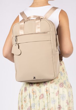 Sac à dos rectangulaire beige avec tirette de fermeture éclair en forme de cœur, porté par une personne vêtue d'un haut blanc et d'une jupe fleurie, vue de dos.