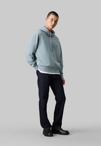 Felpa con cappuccio azzurro chiaro realizzata in cotone, abbinata a jeans blu scuro e mocassini neri. Le caratteristiche notevoli includono una tasca a marsupio frontale.