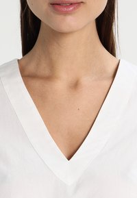 Blouse en lin blanc avec un décolleté en V profond, des manches courtes et un tissu légèrement texturé. Accent sur le détail du décolleté et du col.