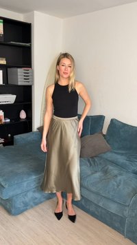 Γυναίκα που στέκεται στο σαλόνι φορώντας μαύρο αμάνικο τοπ, μακριά φούστα από σατέν ελιάς και μαύρα μυτερά τακούνια δίπλα σε ένα μπλε καναπέ.