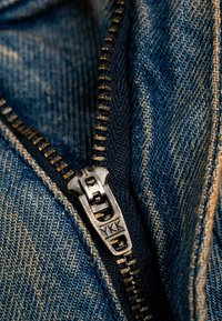 Nahaufnahme eines Metallreißverschlusses auf blauer Denim-Stoff mit sichtbaren Nähten. Der Reißverschluss ist silberfarben und mit "YKK" gekennzeichnet. Die Textur ist glatt und langlebig.