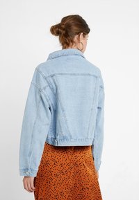 Ljust blå denimjacka med en kortad design, stor krage och synliga sömmar. Har långa ärmar och metallknappar.