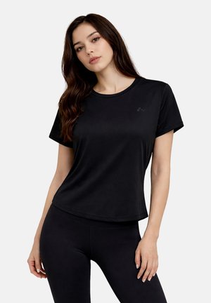 Junge Frau mit langen braunen Haaren, die ein schlichtes schwarzes T-Shirt mit kurzen Ärmeln und schwarze Leggings trägt und vor weißem Hintergrund steht.