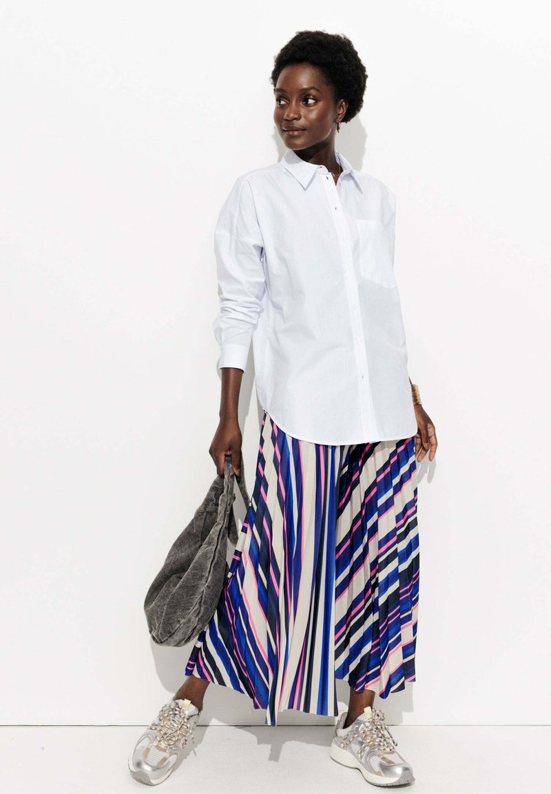 Chemise bleu clair à manches longues, jupe plissée rayée multicolore en bleu, rose et blanc, sac à épaule gris et baskets argentées avec des accents texturés.