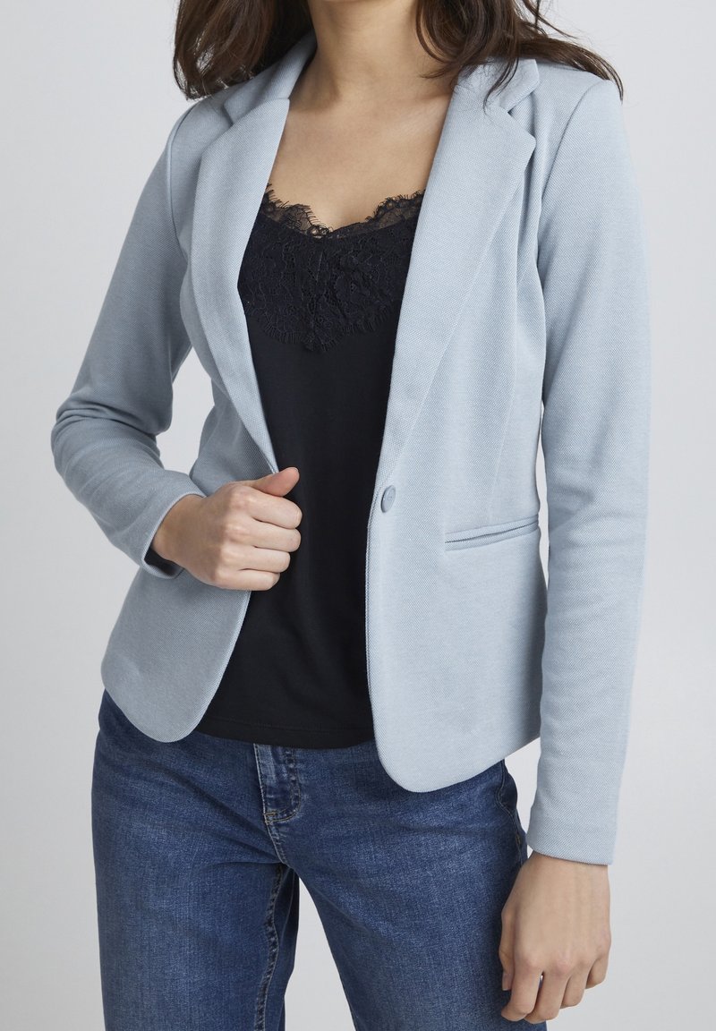 Femme portant un blazer bleu clair sur un haut noir orné de dentelle et un jean bleu, ajustant son blazer d'une main.