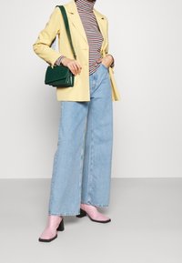 Gelbe Blazer über einem gestreiften Rollkragenpullover, weite, helle blaue Jeans, rosa Ankle Boots und eine grüne strukturierte Handtasche.