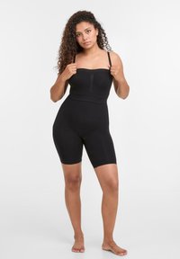 Göndör hajú nő fekete, levehető pántos, pánt nélküli bodysuitot visel, mezítláb áll egy sima, fehér háttér előtt.