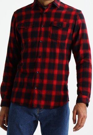 Muž v červeno-černé kostkované košili s dlouhým rukávem, zapínané na knoflíky, se dvěma kapsami na prsou a modrých džínách, stojící na bílém pozadí.