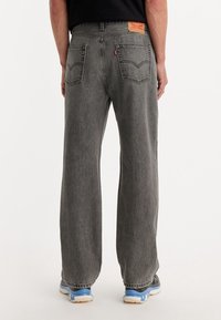 Jean en denim gris à coupe décontractée, avec une étiquette beige et deux poches arrière. L'ourlet inférieur est légèrement effiloché, dévoilant des baskets bleues en dessous.