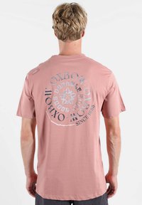 Homme blond portant un t-shirt rose poudré avec un logo circulaire Oxbow et un texte au dos, debout devant un fond blanc uni.