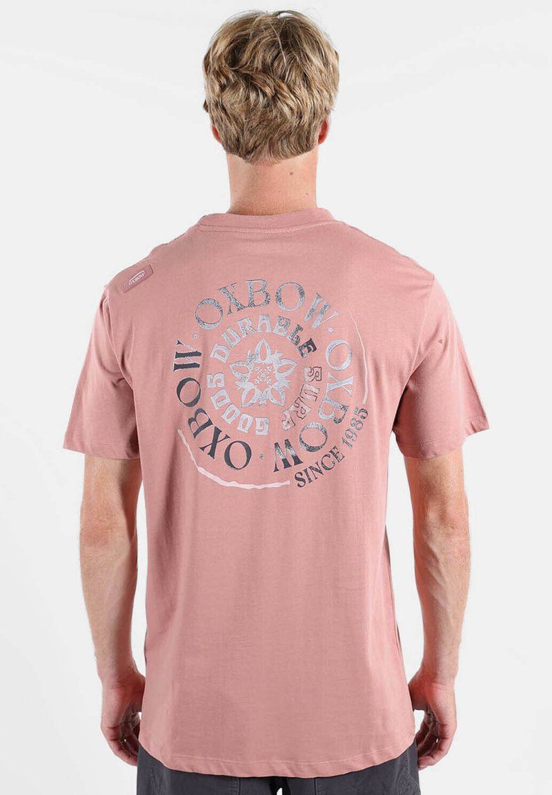 Homme blond portant un t-shirt rose poudré avec un logo circulaire Oxbow et un texte au dos, debout devant un fond blanc uni.
