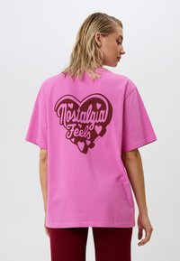 Roza kokvilnas T-kreklu ar lielu brūnu sirds grafiku, uz kuras ir rakstīts "Nostalgia Feels" un nelielas sirds akcenti. Īsas piedurknes un brīvs piegriezums.