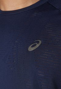 Tumši zils sporta krekls ar teksturētu, perforētu rakstu. Iekļauts atstarojošs logotipa akcenti. Īsas piedurknes un apaļa kakla izgriezums.