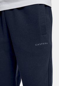 Mørkeblå joggebukser laget av mykt stoff, med en sidelomme og en "CASPARA" logo i lys grå på venstre lår.