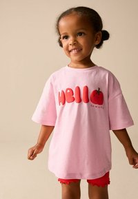 Růžové bavlněné tričko s červeným nápisem "HELLO" a malým motivem rajčete. Design zahrnuje nadměrně velké krátké rukávy a volný střih.