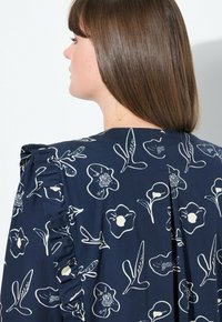 Abito navy con motivo floreale bianco, caratterizzato da spalline arricciate e un dettaglio di cucitura sul retro. Tessuto dalla texture liscia.