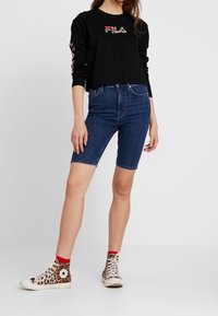 Černá dlouhá košile s logem "FILA" v barevném tisku, spárovaná s modrými džíny a teniskami s leopardím vzorem a červenými akcenty.