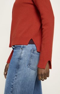 Person in einem roten Pullover mit seitlichen und Bündchenschlitzen, kombinert mit hellblauen Jeans, der Ringe an der Hand trägt.