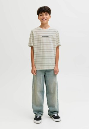 Usměvavý chlapec stojí, má na sobě lehké pruhované tričko, volné vybledlé modré džíny a černé tenisky s bílými podešvemi na jednoduchém pozadí.