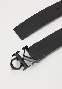 Zwarte leren riem met een matte zwarte metalen gesp met een opvallend logo-ontwerp. De riem heeft een gladde textuur en een elegante, moderne vorm.